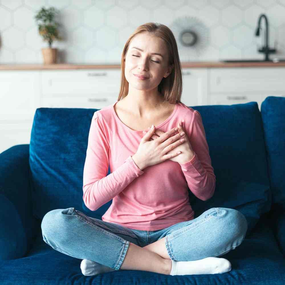 Woman in pink shirt sitting on blue couch with hands on chest, practicing self-compassion in NYC.