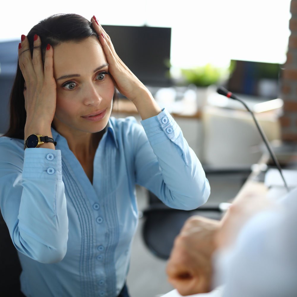 Woman in a blue shirt covering her head with hands in an office, tackling procrastination in NYC.