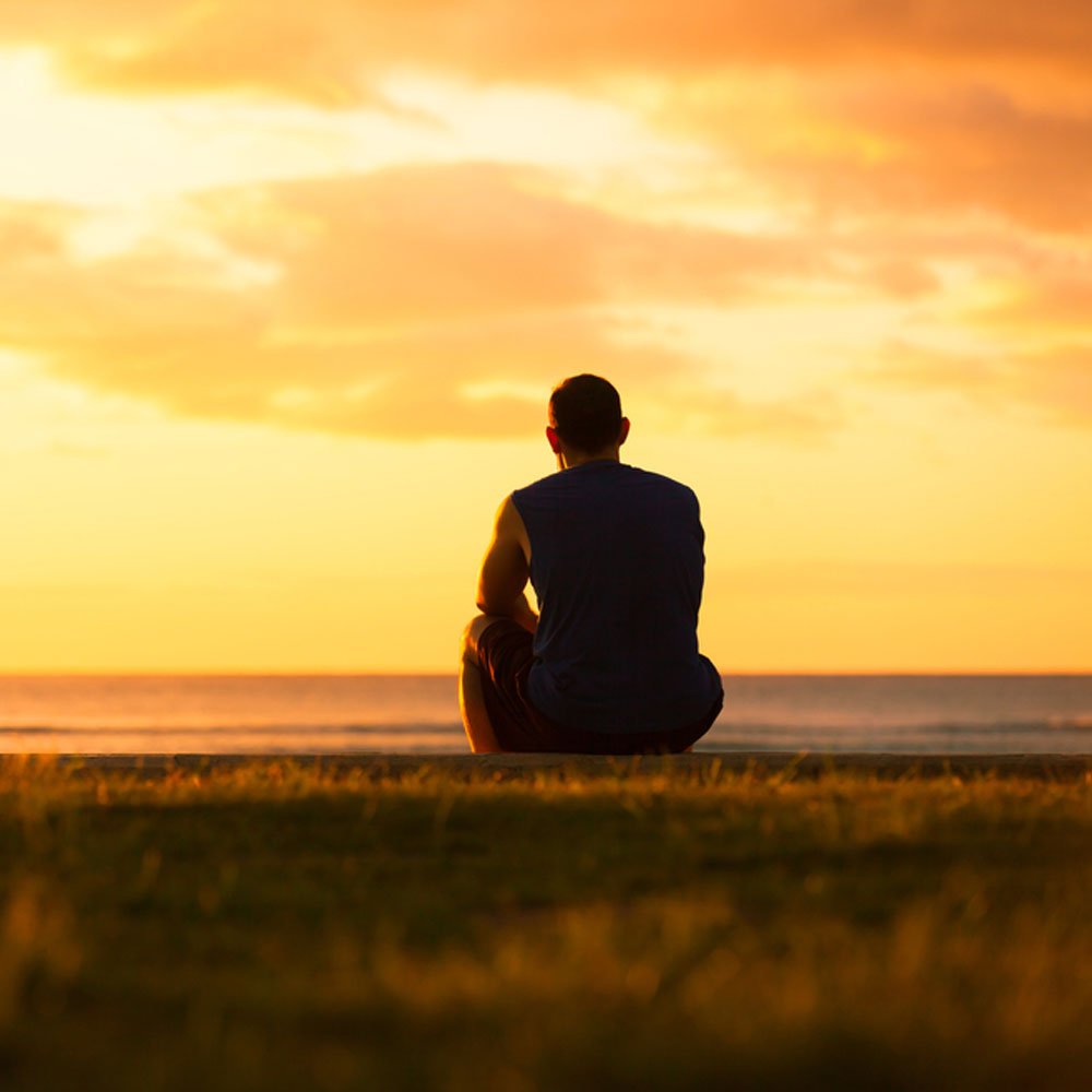 Person sitting on grass facing a sunset, exploring silent suicide in NYC.