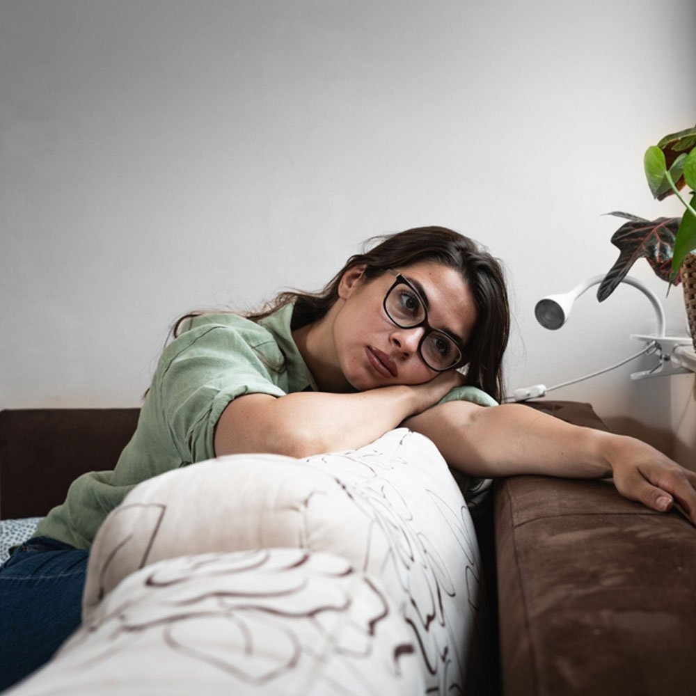 Person resting head on arms on a couch pillow with a plant and lamp, exploring overthinking in NYC.