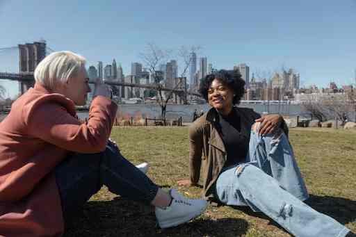 Two people sitting on grass with the Brooklyn Bridge and NYC skyline in the background.
