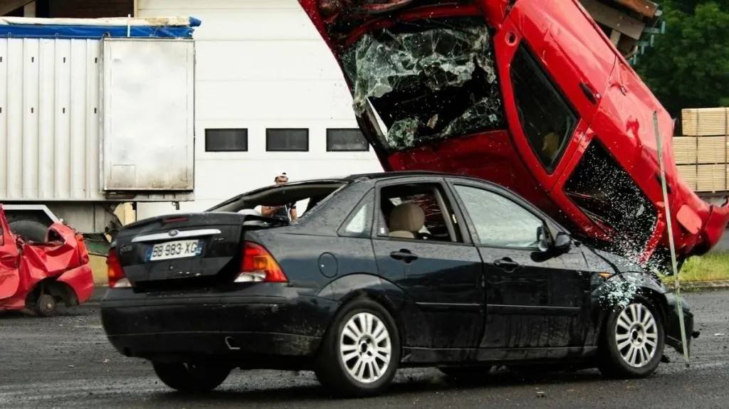 A black car sits on a driveway as a red car is dramatically overturned.